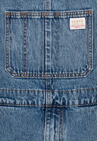 Džinsiniai kombinezonai su mėlynu plovimu, turintys siuvinėtus akcentus, gale esančią kišenę su baltu Levi's logotipo žyme ir tvirta audinio tekstūra.