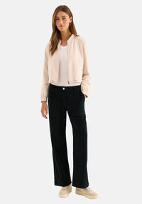 Vrouw die staat in een lichtbeige bomberjack, wit t-shirt, zwarte wijde broek en beige sneakers, tegen een effen achtergrond.
