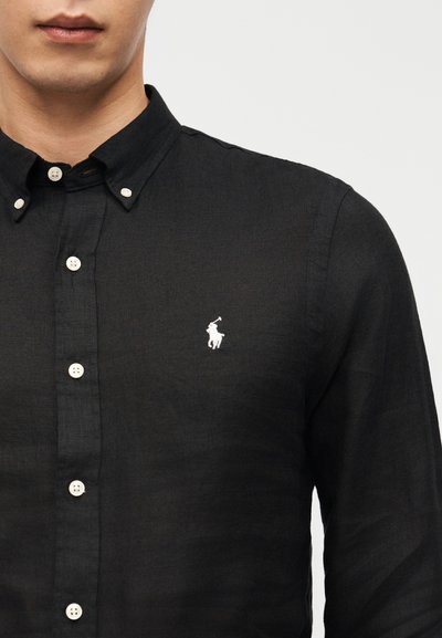 Bărbat purtând o cămașă neagră cu nasturi, având un mic logo brodat alb cu un jucător de polo pe pieptul stâng.