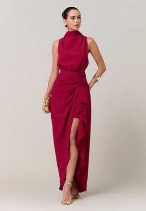 Donna che indossa un abito rosso senza maniche con scollo alto e spacco frontale, accessoriata con braccialetti d'oro, orecchini, pochette e tacchi.