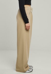 Pantalons beige taille haute en tissu lisse, avec une coupe large et un détail de plis subtil, associés à des baskets grises.