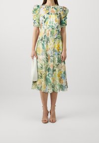 Blumenmidi-Kleid aus leichtem, transparentem Stoff mit Puffärmel, einer Taillennaht und einem gewellten Saum. Kombiniert mit einer weißen Handtasche und Riemchen-Heels.