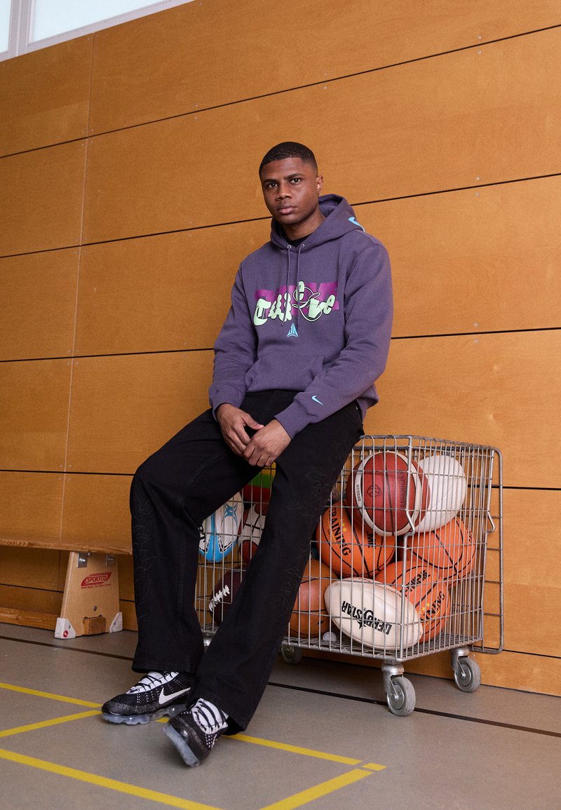 Szara bluza z kolorowym nadrukiem, czarne spodnie i czarne buty z białymi akcentami. Siedzi obok metalowego kosza wypełnionego różnymi piłkami sportowymi.
