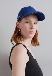 Modrá textilní baseballová čepice s kloboukem a prošívanými detaily. Modelka má na sobě černý bezrukávový top. Hladká textura a ležérní design.