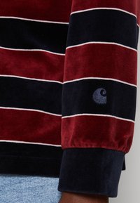 Langemouwenhemd met brede horizontale bordeauxrode en zwarte fluwelen strepen, witte omlijning en een klein geborduurd logo nabij de manchetten.