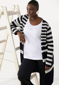 Długi granatowy i biały sweter w paski noszony na białej koszuli z subtelnym haftem, czarne spodnie i duże okrągłe kolczyki. Opierająca się na drabinie.