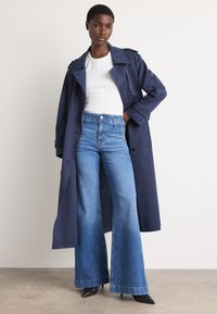 Manteau trench marine avec épaulettes, haut ajusté blanc et Jean bleu à taille haute évasé. Des chaussures à talons pointus noires complètent la tenue.