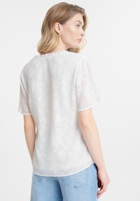 Blusa blanca y transparente con un sutil patrón floral, mangas cortas y cuello con volantes, combinada con jeans de mezclilla azul claro.