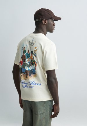 Férfi krémszínű pólóban, színes illusztrációval, amely embereket ábrázol, amint kenyérrel osztoznak egy asztal körül, a hátoldalon a "Silling Pieces Breaking Bread" felirattal.