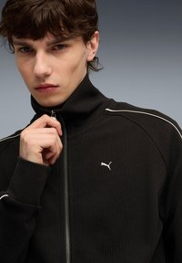 Zwarte zip-up jack met hoge kraag, textuurstof, witte accenten op de schouders en een klein wit logo op de borst.