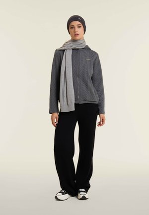 Giacca grigia con zip e trama a trecce, abbinata a una sciarpa grigia chiara, pantaloni neri a gamba larga e sneakers bianche e nere.