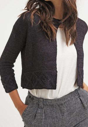 Femme portant un cardigan noir court en maille avec des motifs géométriques sur les poignets et l’ourlet, un haut blanc et un pantalon gris à carreaux taille haute.