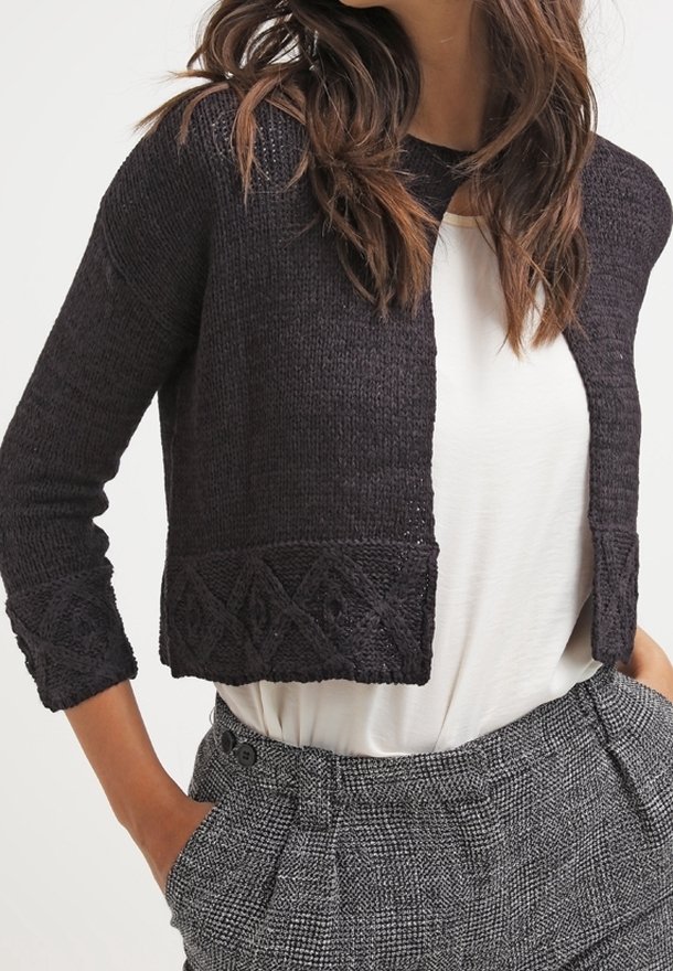 Femme portant un cardigan noir court en maille avec des motifs géométriques sur les poignets et l’ourlet, un haut blanc et un pantalon gris à carreaux taille haute.