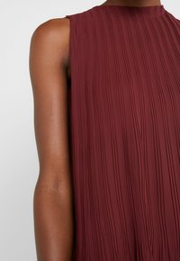 Mörkröd ärmlös klänning med vertikala veck, hög halsringning och textilstruktur. Klänningen har en avslappnad passform och faller elegant.
