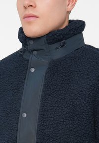 Muškarac nosi tamnoplavu jaknu od teksturirane flis tkanine s dugmadi na visokom ovratniku i špulicama za podešavanje na bijeloj pozadini.