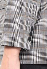 Manica di un blazer a quadri grigio e nero con una texture liscia, caratterizzata da tre bottoni neri e linee arancioni a contrasto nel motivo.