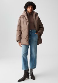 Giacca a vento marrone con colletto alto, abbinata a jeans a gamba larga di un blu chiaro e stivaletti neri con punta appuntita. Texture liscia in tutto il capo.
