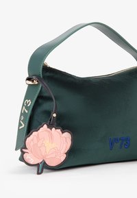 Groene fluwelen handtas met een gladde textuur, voorzien van een gouden rits, een blauw logo en een roze bloemencharme voor detail.