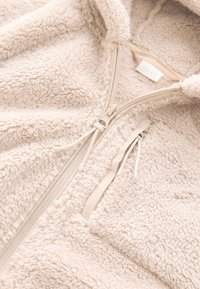 Jachetă crem din fleece cu fermoar, cu suprafață moale și texturată, având un fermoar frontal și șnururi ajustabile. Eticheta interioară poartă inscripția "NEXT."