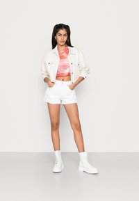 Casaco de ganga curto branco por cima de uma blusa rosa com padrão ondulado e calções brancos com franjas. A modelo usa botins brancos até ao tornozelo com atacadores.