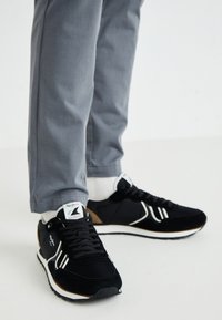 Μαύρα και σουέτ αθλητικά παπούτσια με λευκές λεπτομέρειε�ς, με υφή πλέγματος και καουτσούκ σόλα. Γκρι παντελόνι μερικώς ορατό.