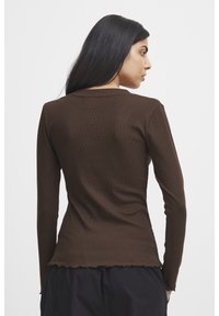 Braun, langärmliges, geripptes Oberteil mit rundem Halsausschnitt und gewelltem Saum, getragen von einem Model, das von hinten schaut, und das den figurbetonten Schnitt und den strukturierten Stoff zeigt.