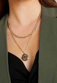 Collana a catena a più strati tono oro, caratterizzata da anelli texturizzati e un pendente circolare con dettagli intricati al centro.