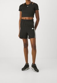 Black cropped athletic top with cut-out details, paired with black shorts featuring white side stripes and logo. Black athletic shoes complete the look.