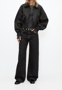 Veste noire courte avec col montant, manches évasées, associée à un jean noir taille haute à jambes larges avec des accessoires contrastants.