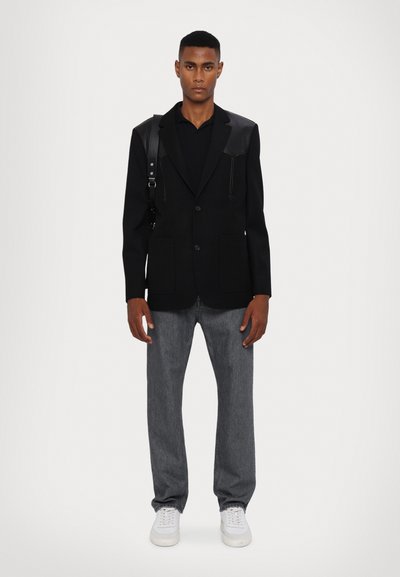 Zwarte blazer met leren accenten, gecombineerd met een zwart shirt en grijze jeans. Witte sneakers maken de outfit compleet. Model staat tegen een neutrale achtergrond.