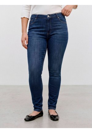 Person wearing dark blue skinny jeans and black ballet flats standing on a light gray floor against a plain background.
