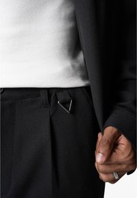 Zwarte broek van een gestructureerde stof, met een metalen driehoekige clip op de tailleband en een witte geribde shirt zichtbaar eronder.