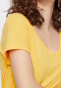 Person med lockigt brunt hår som bär en gul kortärmad V-halströja, som visar övre delen av kroppen och nacken.