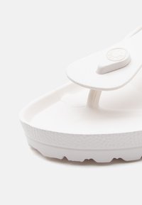 Κοντινό πλάνο λευκού σαγιονάρου με υφή, τρίγωνο λουράκι και ανάγλυφο κυκλικό λογότυπο στο λουράκι.