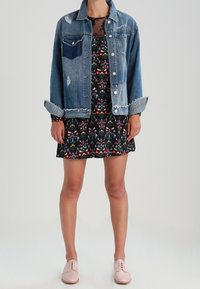 Mujer con una chaqueta de mezclilla azul sobre un vestido negro con estampado floral, combinada con zapatos oxford rosas claros, de pie frente a un fondo liso.