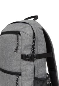 Gray fabric backpack with black zippers and pull cords, side compression strap with buckle, padded shoulder strap, and mesh side pocket.