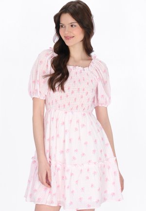 Młoda kobieta o długich brązowych włosach nosi różową sukienkę w kwiaty z krótkim rękawem, marszczonym gorsem i falbanianą spódnicą, spogląda w bok i uśmiecha się.