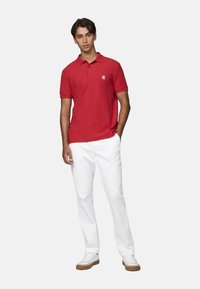 Camisa polo roja con cuello y mangas cortas, combinada con pantalones blancos. La camisa presenta un pequeño logo en el lado izquierdo del pecho.