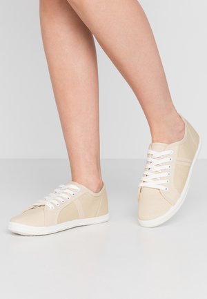 Nogi w beżowych tenisówkach z płótna z białymi sznurowadłami i białymi podeszwami, jedna stopa lekko uniesiona na jednolitym tle.