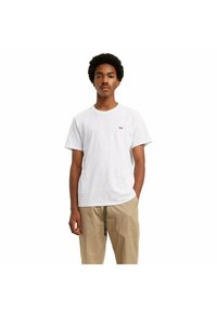 Camiseta blanca de algodón con un pequeño logo rojo de Levi's en el lado izquierdo del pecho, combinada con pantalones beige. Diseño de color sólido y cuello redondo.