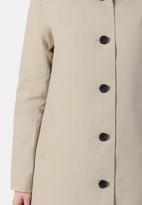 Cappotto beige con taglio dritto, bottoni neri, tasche laterali e tessuto dalla consistenza liscia. Adatto a vari contesti casual.