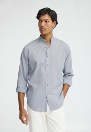 Uomo che indossa una camicia azzurro chiaro con collo alla coreana e maniche rimboccate, e pantaloni bianchi, guarda di lato su uno sfondo neutro.