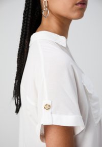 Camicia bianca a maniche corte con colletto, caratterizzata da dettagli ai polsini con bottoni. Tessuto liscio con una leggera lucentezza. Orecchini a bottone visibili nell'immagine.