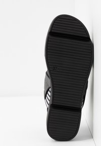 Semelle en caoutchouc noir d'une chaussure avec une adhérence texturée, numéro de taille 37 visible sur la semelle, reposant verticalement contre des surfaces blanches.