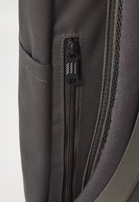 Détail d'un sac à dos en tissu gris montrant une fermeture éclair et une poche latérale avec un tirant noir. Comprend des coutures et une doublure intérieure en maille.