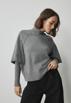 Frau trägt einen grauen, geschichteten Strickpullover mit gerippten Ärmeln und Ausschnitt, kombiniert mit hoch taillierter schwarzer Hose, posiert vor hellem Hintergrund.