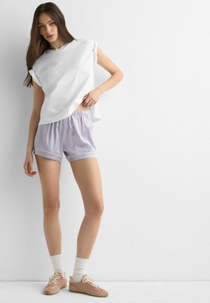 Femme debout contre un mur blanc, portant un haut blanc sans manches, un short violet clair, des chaussettes blanches et des baskets roses.