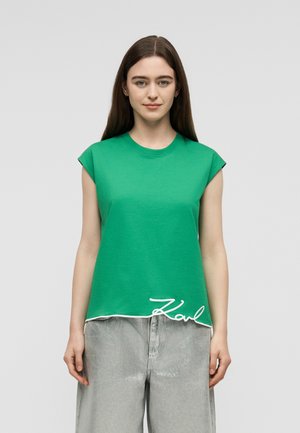 Jeune femme aux longs cheveux bruns portant un haut vert sans manches avec des bordures blanches et un pantalon gris ample sur un fond gris uni.