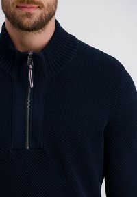 Donkerblauwe gebreide pullover met hoge kraag en kwart-rits aan de voorkant. Geprofileerde stof en contrasterende ritshendel met accenten in rood en wit.
