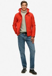Veste à fermeture éclair rouge avec capuche, pull gris texturé, jean bleu et chaussures noires. Coupe slim avec une poche poitrine sur la veste.